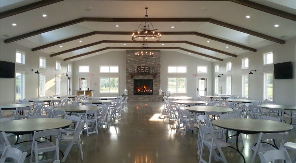 Large event hall featuring a central stone fireplace, high ceilings with exposed beams, round tables, and ample natural light.