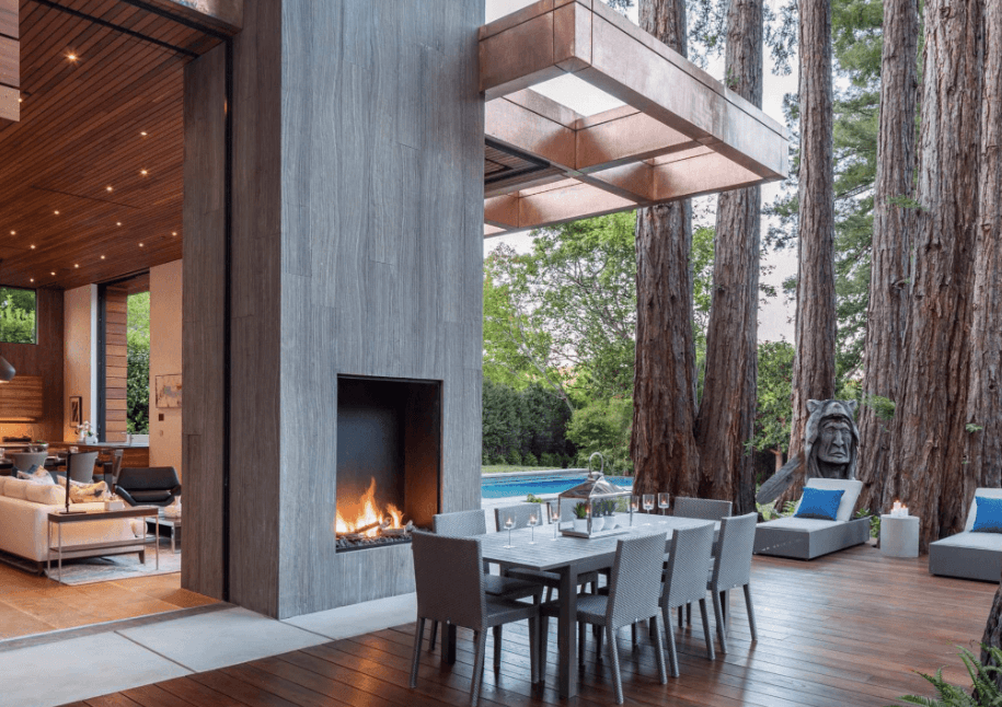 Large outdoor patio surrounded by tall trees with an open outdoor fireplace in the middle