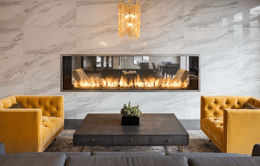 Gray couch and yellow chairs placed around a coffee table with a marble wall featuring a see-through linear fireplace in the background