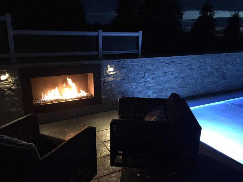 A modern, single-sided outdoor gas fireplace set into a stone wall, glowing warmly with flames at night