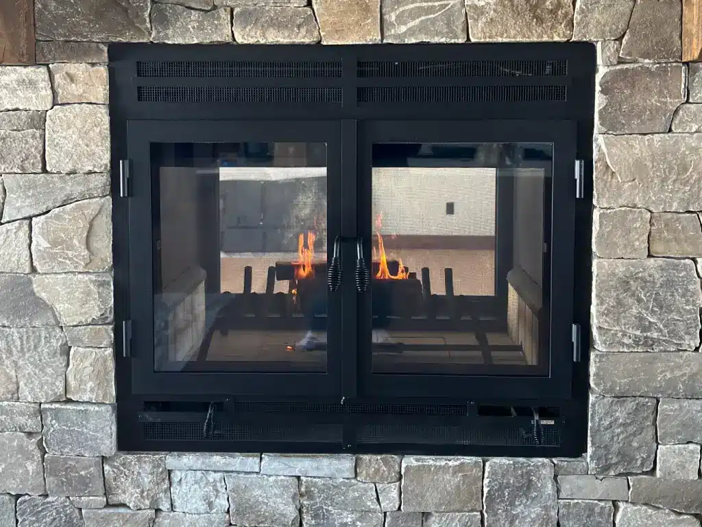 Acucraft Hearthroom 44 see-through wood fireplace burning behind closed glass doors in a stone wall.